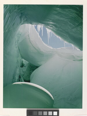 A color photograph from inside of a blue and green ice cave showing a sliver of blue sky.