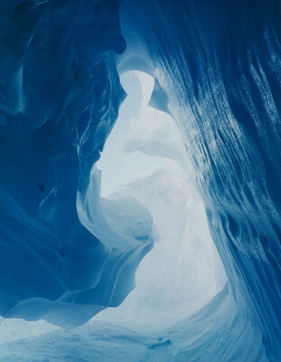 A color photograph taken inside an ice cave with swirling blue walls and a bright opening toward daylight.