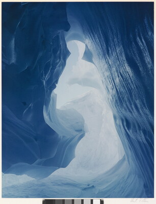 A color photograph taken inside an ice cave with swirling blue walls and a bright opening toward daylight.