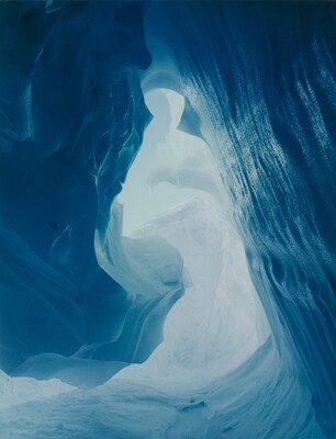 A color photograph taken inside an ice cave with swirling blue walls and a bright opening toward daylight.