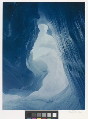 A color photograph taken inside an ice cave with swirling blue walls and a bright opening toward daylight.