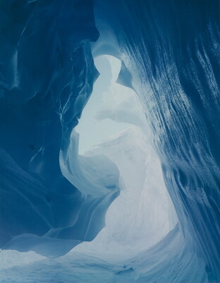A color photograph taken inside an ice cave with swirling blue walls and a bright opening toward daylight.