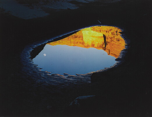 A color photograph of a pool of water reflecting a clear blue sky with a moon and orange mountain cliffs.
