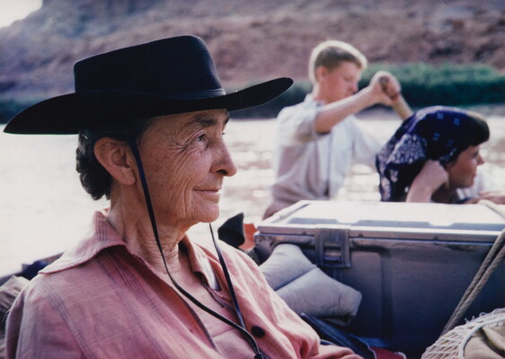 A color photograph of an older White woman in profile wearing a black gaucho hat sitting in a boat on a river.