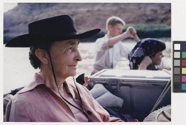 A color photograph of an older White woman in profile wearing a black gaucho hat sitting in a boat on a river.
