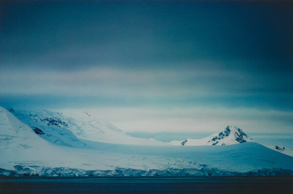 A color photograph of a snowy coastline with mountains and dense clouds.