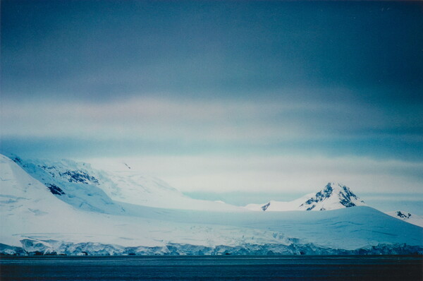 A color photograph of a snowy coastline with mountains and dense clouds.