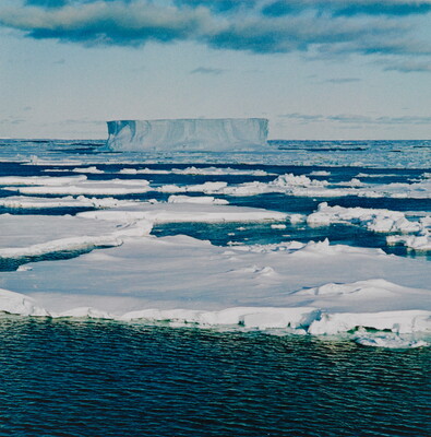 A color photograph of an ocean covered with ice floes and a large, flat-topped iceberg in the distance.