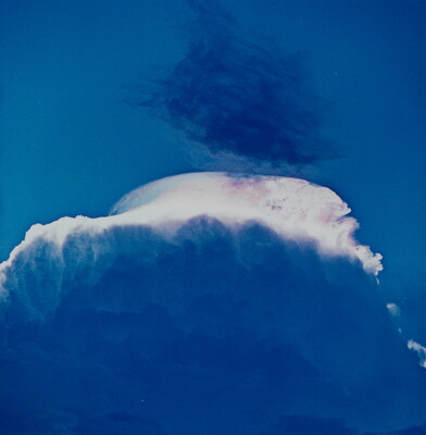 A color photograph of a large storm cloud with a striking white top and dark blue base.
