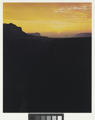 A color photograph of dark black and purple canyon walls in silhouette beneath a bright yellow and orange sunrise.