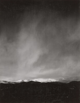 A black-and-white photograph of a snow-covered mountain range with a dark storm forming in the sky.