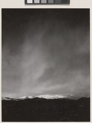 A black-and-white photograph of a snow-covered mountain range with a dark storm forming in the sky.