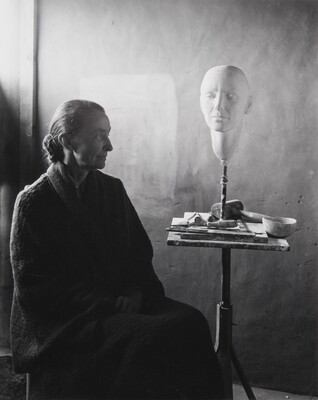 A black-and-white portrait photograph of a seated older White woman wearing a dark, floor length dress; a white face mask sits on a stand on a cluttered end table.
