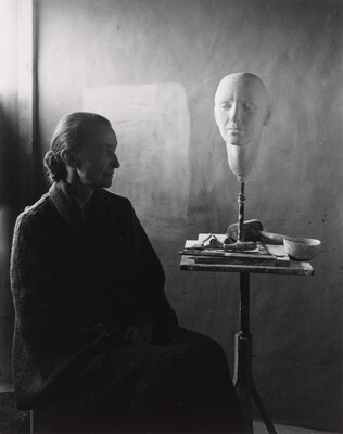 A black-and-white portrait photograph of a seated older White woman wearing a dark, floor length dress; a white face mask sits on a stand on a cluttered end table.