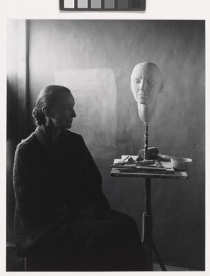 A black-and-white portrait photograph of a seated older White woman wearing a dark, floor length dress; a white face mask sits on a stand on a cluttered end table.