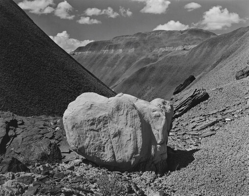 A black-and-white photograph of a light-colored boulder sitting in scree with dark mountains in the background.