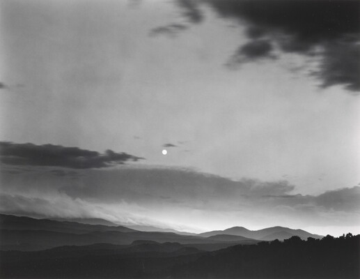 A black-and-white photograph of a mountainous landscape with a bright white moon in a cloudy sky.