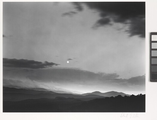 A black-and-white photograph of a mountainous landscape with a bright white moon in a cloudy sky.