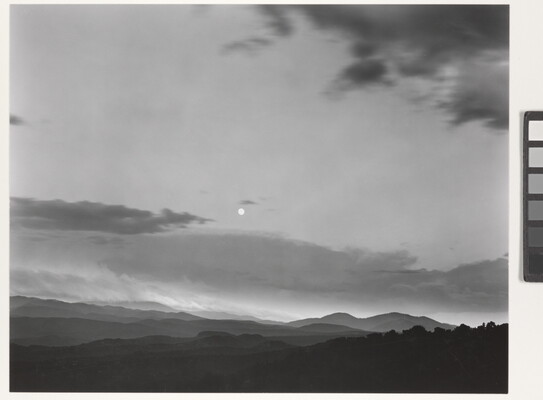 A black-and-white photograph of a mountainous landscape with a bright white moon in a cloudy sky.
