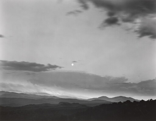 A black-and-white photograph of a mountainous landscape with a bright white moon in a cloudy sky.