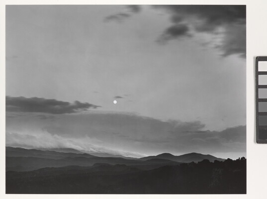 A black-and-white photograph of a mountainous landscape with a bright white moon in a cloudy sky.