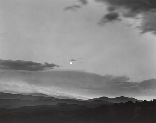 A black-and-white photograph of a mountainous landscape with a bright white moon in a cloudy sky.