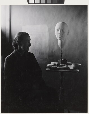 A black-and-white portrait photograph of a seated older White woman wearing a dark, floor length dress; a white face mask sits on a stand on a cluttered end table.