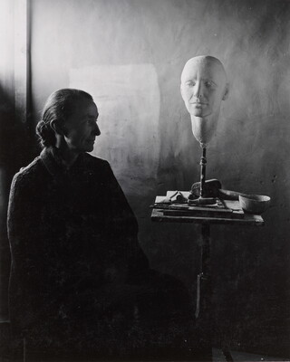 A black-and-white portrait photograph of a seated older White woman wearing a dark, floor length dress; a white face mask sits on a stand on a cluttered end table.