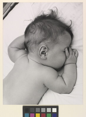A black-and-white photograph of a White baby with dark hair lying on his stomach, thumb of one hand in his mouth, on white sheets.