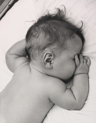 A black-and-white photograph of a White baby with dark hair lying on his stomach, thumb of one hand in his mouth, on white sheets.