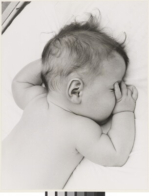 A black-and-white photograph of a White baby with dark hair lying on his stomach, thumb of one hand in his mouth, on white sheets.
