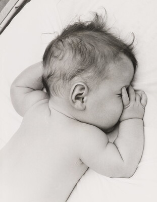 A black-and-white photograph of a White baby with dark hair lying on his stomach, thumb of one hand in his mouth, on white sheets.