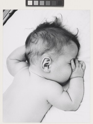 A black-and-white photograph of a White baby with dark hair lying on his stomach, thumb of one hand in his mouth, on white sheets.
