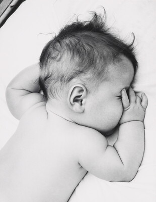 A black-and-white photograph of a White baby with dark hair lying on his stomach, thumb of one hand in his mouth, on white sheets.