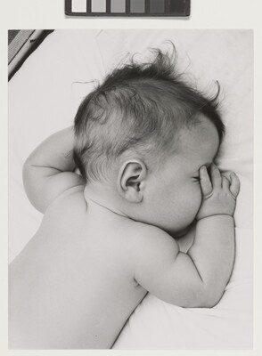 A black-and-white photograph of a White baby with dark hair lying on his stomach, thumb of one hand in his mouth, on white sheets.