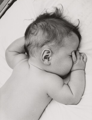 A black-and-white photograph of a White baby with dark hair lying on his stomach, thumb of one hand in his mouth, on white sheets.