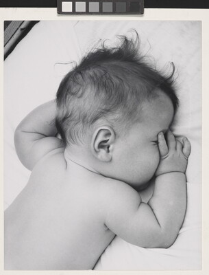 A black-and-white photograph of a White baby with dark hair lying on his stomach, thumb of one hand in his mouth, on white sheets.