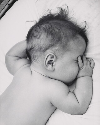 A black-and-white photograph of a White baby with dark hair lying on his stomach, thumb of one hand in his mouth, on white sheets.