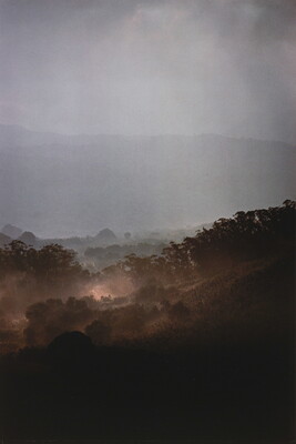 A color photograph of a hazy rain falling on a tree-filled, hilly landscape.