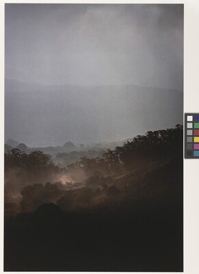 A color photograph of a hazy rain falling on a tree-filled, hilly landscape.