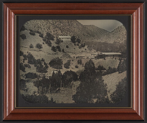 A black-and-white photograph of several buildings on a mountainside dotted with pine trees.