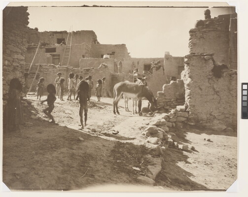 A black-and-white photograph of Indigenous children playing in a courtyard surrounded by a multi-level pueblo as donkeys graze nearby.