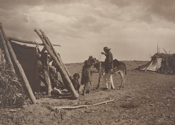 A black-and-white photograph of Indigenous people, two adults and a child, tacking up a donkey near several wood and grass structures.