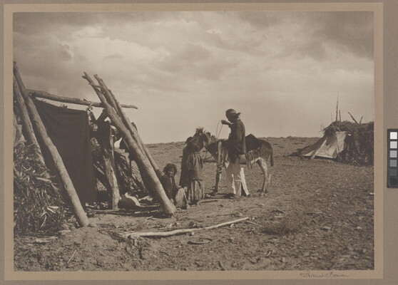 A black-and-white photograph of Indigenous people, two adults and a child, tacking up a donkey near several wood and grass structures.