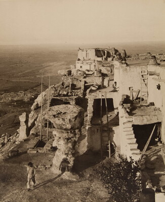 A black-and-white photograph of a multi-level pueblo building on top of a mesa, as viewed from above.