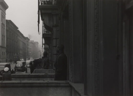 A black-and-white photograph of rows of buildings and townhomes on a city street; two Black men stand on neighboring stoops looking toward the street.