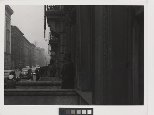 A black-and-white photograph of rows of buildings and townhomes on a city street; two Black men stand on neighboring stoops looking toward the street.