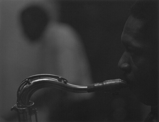 A black-and-white close-cropped photograph of a Black man in profile playing a saxophone.