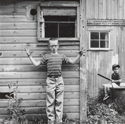 A black-and-white photograph of a White boy with a buzz cut, glasses, wearing a striped shirt standing in front of a wood building with his arms wide and fingers splayed; another child is seated on a wood box in the background.