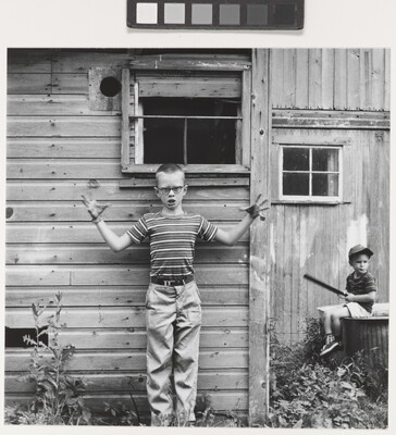 A black-and-white photograph of a White boy with a buzz cut, glasses, wearing a striped shirt standing in front of a wood building with his arms wide and fingers splayed; another child is seated on a wood box in the background.